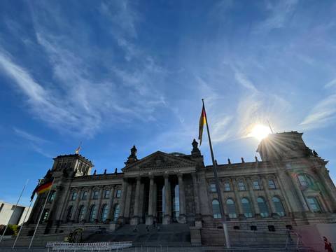       Reichstag building under a bright blue sky with the sun flaring beside the German flag.
  