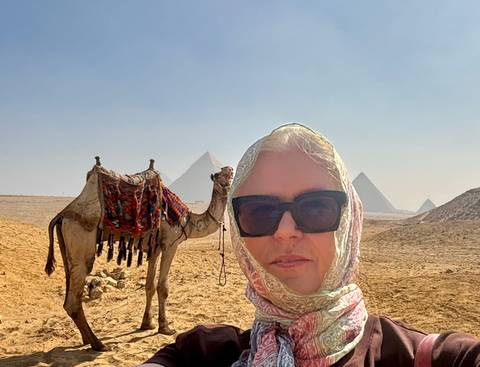       Selfie of a woman in headscarf and sunglasses with a camel and the Pyramids of Giza in the hazy background.
  