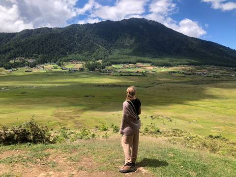       A young woman wrapped in a shawl gazes across the wide green valley floor of Phobjikha with forested hills rising behind.
  