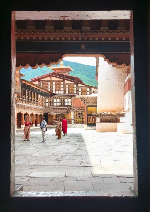       Visitors explore the ornate courtyard of a traditional Bhutanese monastery with intricate woodwork and mountain scenery beyond.
  