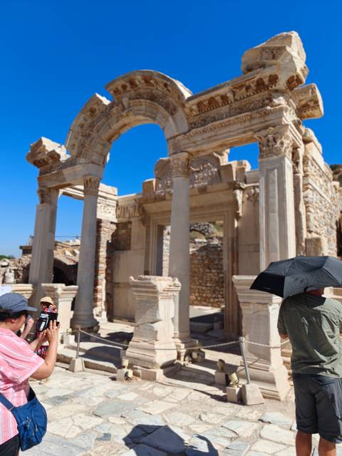       Visitors walk among ornate stone arches and columns of an ancient Roman gate under bright blue sky.
  