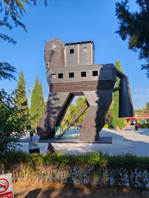       Large wooden replica of the Trojan Horse towers above visitors in a leafy park.
  