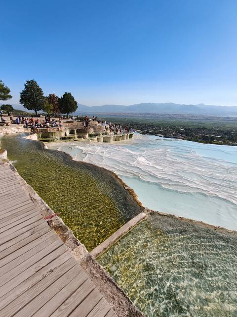       White travertine terraces overflow with turquoise water overlooking a valley filled with visitors.
  