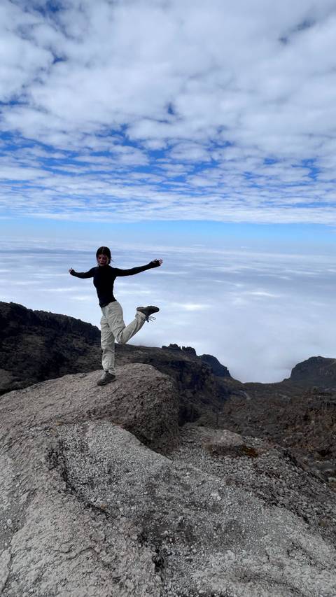       Female trekker balances playfully on a rocky outcrop high above the cloud layer on Kilimanjaro.
  