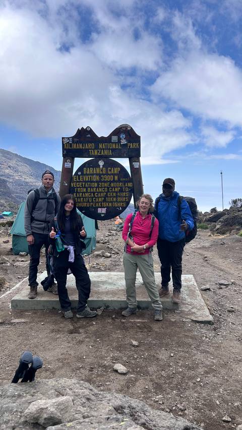       Trekkers stand at the Barranco Camp sign on Kilimanjaro with tents and rocky slopes behind.
  