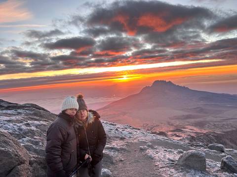       Two climbers wrapped up warm watch a dramatic sunrise over Kilimanjaro's peaks with frosted ground.
  