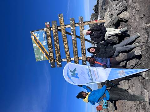       Summit team celebrates at Uhuru Peak sign under clear blue skies on top of Kilimanjaro.
  