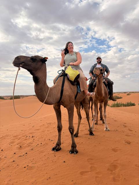       Two travelers ride camels across rolling orange sand dunes, a cloudy sky overhead.
  