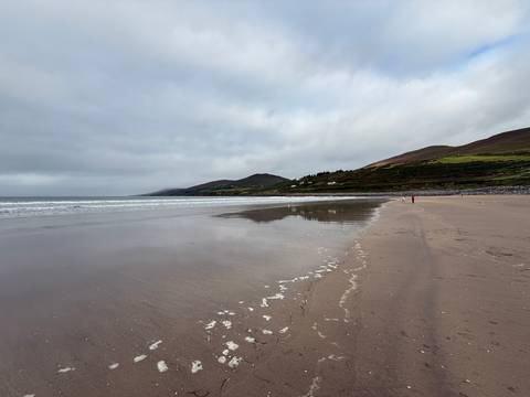       Wide, empty sandy beach with gentle surf and distant green hills beneath a grey, cloud-filled sky.
  