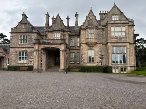       Grand Victorian manor house with stone façade, arched portico entrance and manicured hedges.
  