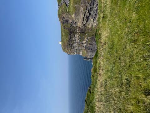       Steep grassy cliff jutting into a calm blue Atlantic with a small white building perched on top.
  