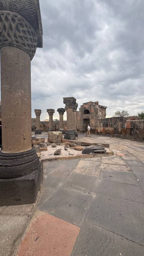       Traveler exploring the columned ruins of Zvartnots Cathedral under a cloudy sky.
  