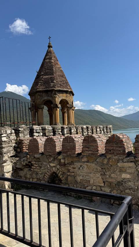       Stone bell tower of a medieval fortress overlooking a turquoise reservoir and forested mountains.
  