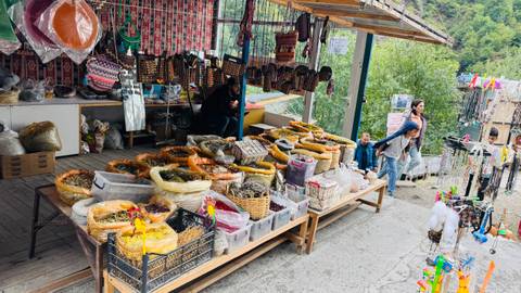       Bustling roadside stall selling baskets of dried fruit, honey rings and souvenirs near a forested area.
  