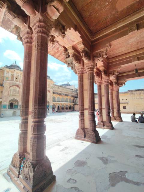       Sun-lit courtyard of Amber Fort framed by ornate sandstone pillars with tourists exploring the space.
  