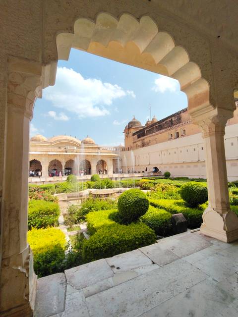       Lush garden courtyard with fountains framed by palace arches under a bright sky.
  