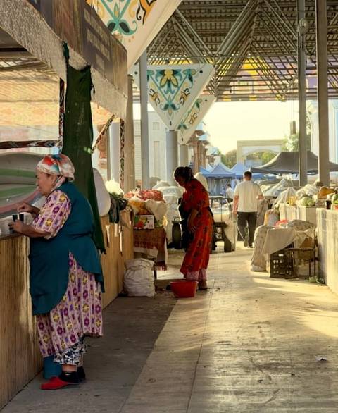       Covered bazaar aisle with vendors arranging produce and fabrics in warm afternoon light.
  