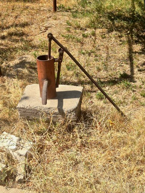       Old rusty hand water pump fixed on a concrete block in a dry grassy field.
  