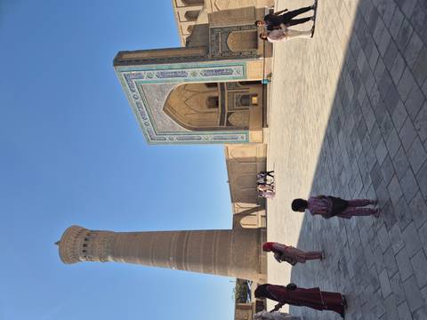       Spacious courtyard of an ancient madrasah with towering minaret and patterned blue-tile portal, visitors strolling across the stone floor.
  