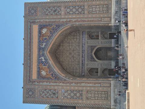       Grand facade of Sher-Dor Madrasah with intricate mosaic and visitors gathered on the steps.
  