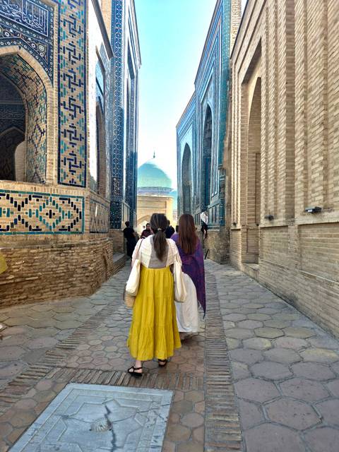      Narrow tiled alleyway in Samarkand with tourists walking toward a distant turquoise dome.
  
