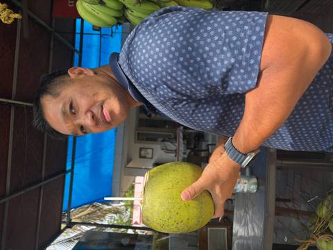       Traveler holding a fresh coconut with a straw at an open-air café under a blue tarp roof.
  