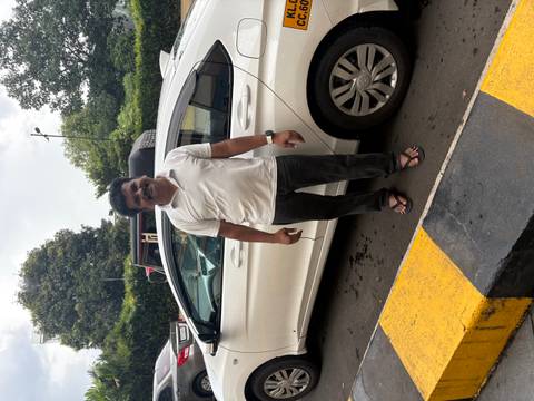       Driver standing beside a white sedan taxi in a parking area on an overcast day.
  
