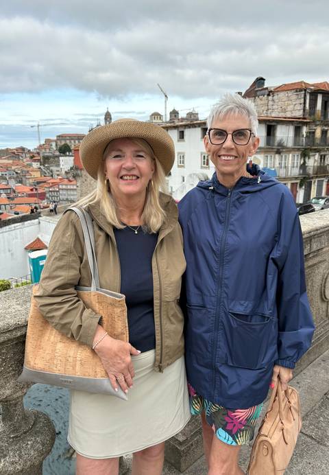       Two women in hats and jackets pose with Porto’s tiled rooftops and Douro riverside district behind them on an overcast day.
  