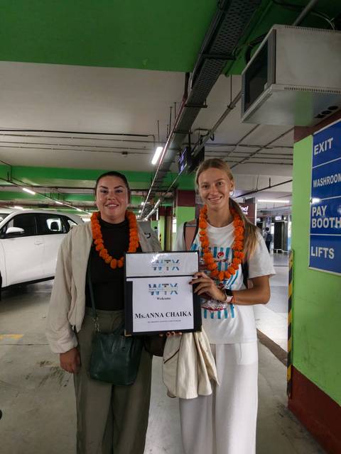       Two women wearing marigold garlands holding a welcome sign in an airport parking garage.
  