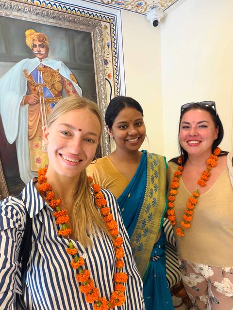       Close selfie of three women wearing marigold garlands and smiling indoors.
  