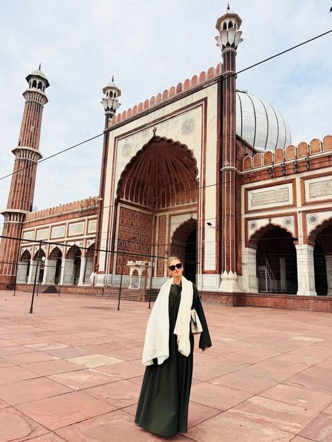       A woman stands in front of the grand arched entrance of Jama Masjid with intricate red-and-white stonework.
  