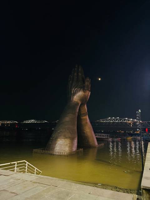       Massive bronzed praying hand sculpture beside a moonlit river with bridges illuminated in the distance.
  