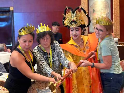       Tourists wearing gold paper crowns interact with a costumed performer in ornate traditional Chinese attire at a banquet.
  
