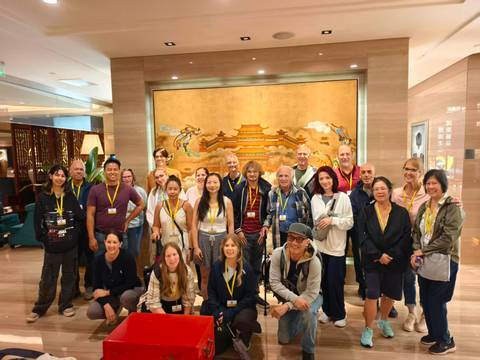       Large tour group poses indoors before a gilded mural depicting an ancient Chinese palace.
  