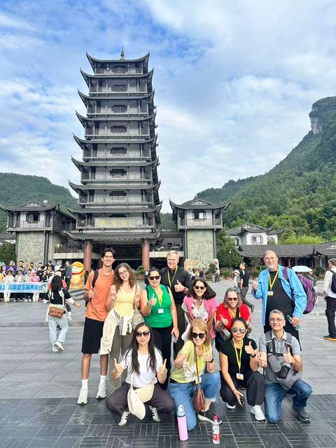       Smiling tourists stand before a multi-story pagoda gate backed by forested mountains.
  