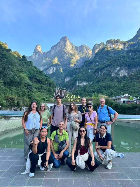       Travel group poses on a bridge beneath dramatic karst peaks and an arched entrance reading Tianmen Mountain.
  
