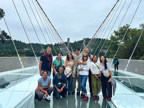       Visitors stand on a glass suspension bridge with cables stretching overhead and forested hills beyond.
  