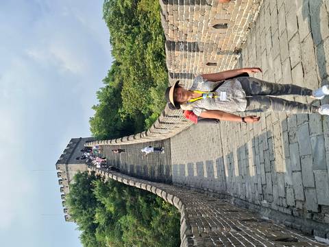       Traveler stands on a stone walkway ascending the Great Wall of China surrounded by green trees and blue sky.
  