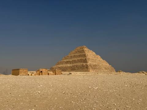       The Step Pyramid of Saqqara stands against a stark, hazy sky with small ruins at its base.
  
