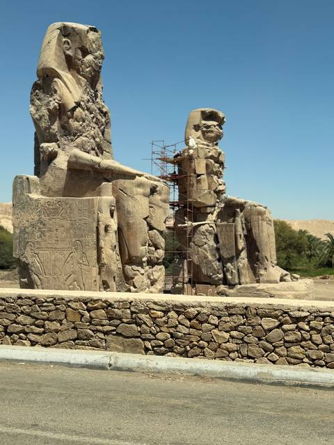       Weathered statues of the Colossi of Memnon with scaffolding amid arid surroundings.
  