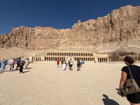       Wide courtyard view of Hatshepsut Temple with visitors walking toward its grand colonnades beneath towering cliffs.
  