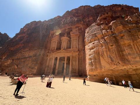       Sunlight streams onto the Treasury’s ornate façade at Petra as visitors explore the sandy plaza.
  