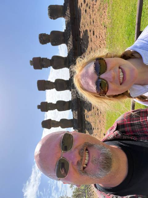       Partial selfie of a couple with a distant row of moai statues under bright daylight.
  