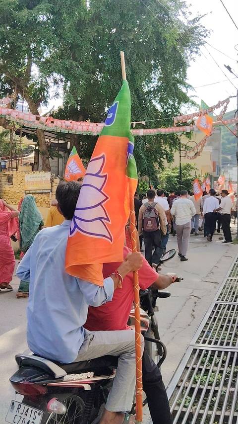       Street procession with people carrying orange political flags featuring a lotus symbol.
  