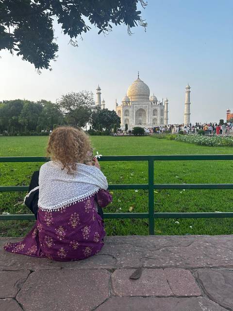       A child wrapped in a shawl gazes at the Taj Mahal from behind a green railing across a lush lawn.
  