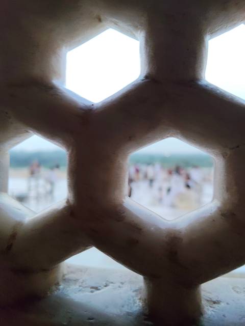       View through a hexagonal marble lattice with blurred crowd and river beyond.
  