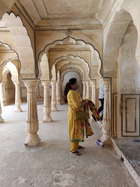       Woman in yellow saree admires a hall of carved sandstone columns and arches.
  