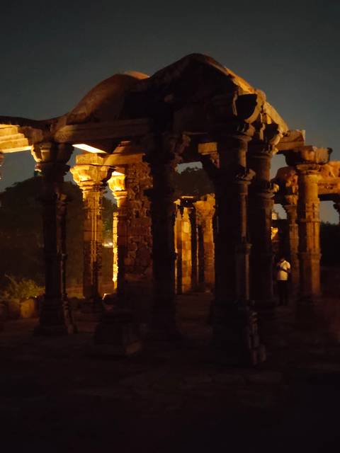       Dimly lit stone columns of an ancient ruin illuminated at night with a visitor in the shadows.
  