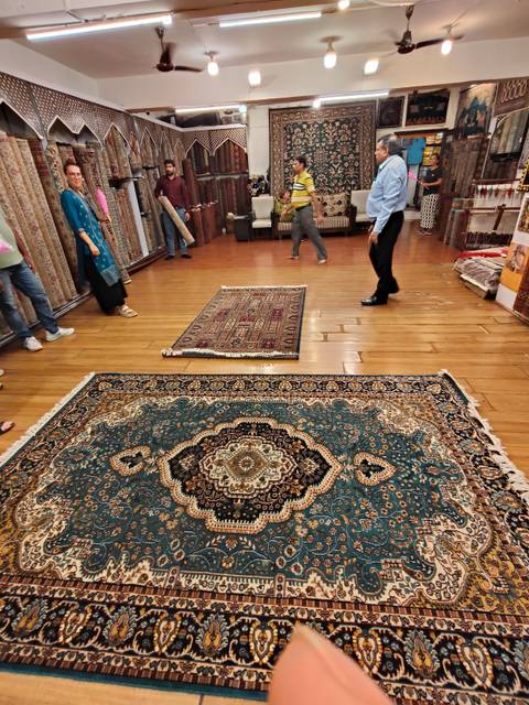       Interior showroom where visitors inspect ornate Persian carpets displayed on polished wooden floors.
  
