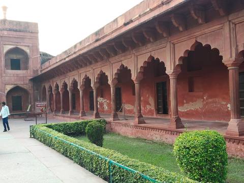       Long red-stone arcade with scalloped arches inside a Mughal complex with a lone visitor.
  